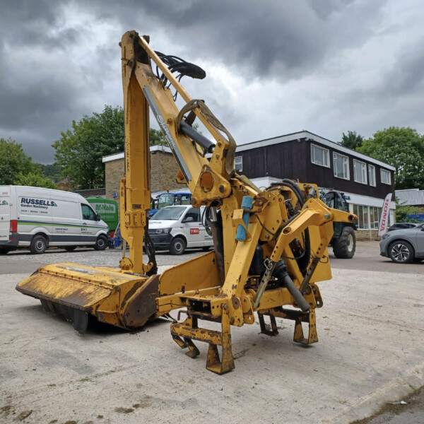 Large yellow construction machine with hydraulic arm and bucket in a parking lot beside vans under dark stormy clouds.