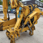 Close-up of yellow construction machinery ( excavator arms and hydraulic cylinders ) in a yard with vans in the background, showing weathered paint and grime for an equipment display.