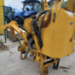 Yellow excavator in the foreground with hydraulic hoses, arms, and tracked feet; blue tractors parked behind a green fence in an outdoor lot.