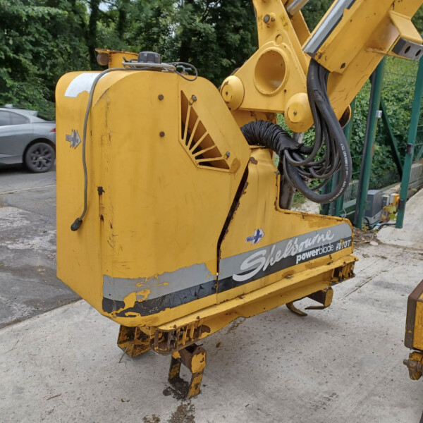 Close-up of a yellow hydraulic construction machine with hoses and metal arms on a construction site. A green fence and trees are in the background, and a car passes by on the road nearby.