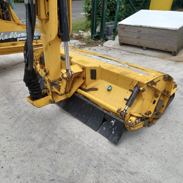 Close-up of a yellow construction excavator blade and hydraulic arm on a concrete surface, with a wooden storage box and an orange traffic cone in the background.