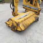 Close-up of a yellow construction machine’s bucket and hydraulic arm resting on a concrete surface, showing wear and rust patches.