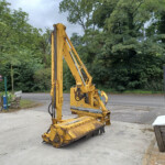 Yellow construction excavator with extended arm on a paved site, surrounded by trees and a green fence in the background.