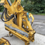 Close-up of a yellow hydraulic construction machine with hoses and a worn attachment, parked on a paved lot with green trees in the background.
