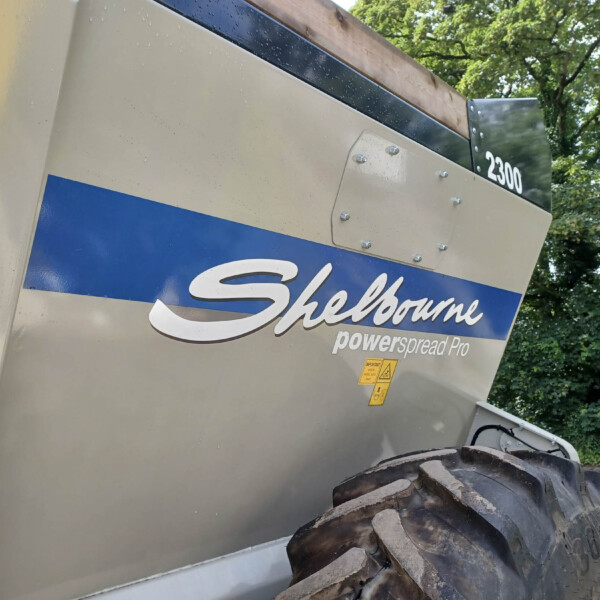 Side view of a large beige and blue Shelbourne Powerspread Pro vehicle, with a large tire in the foreground and green trees in the background.