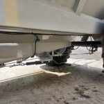 Underside of a large industrial trailer showing black cables, green connectors, metal beams, and a wheel on dirt ground surrounding it outside.