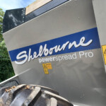 Close-up of a gray industrial trailer with a blue Shelbourne sign and several yellow warning labels; a tipped dumpster filled with logs sits in front, and green foliage is in the background.