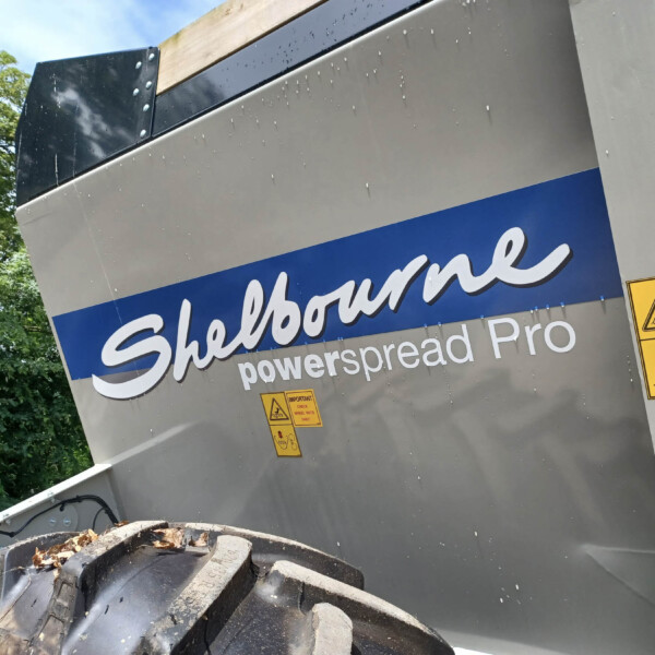 Close-up of a gray industrial trailer with a blue Shelbourne sign and several yellow warning labels; a tipped dumpster filled with logs sits in front, and green foliage is in the background.