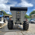 Rear view of a large gray grain trailer with a black top, big off-road tires, and a metal ladder, in a farm yard with a blue tractor nearby.