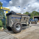 New Holland Agriculture sign above a yard with large farm machinery and a trailer, blue sky and trees in the background