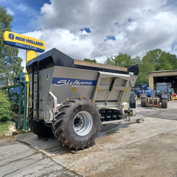 New Holland Agriculture sign above a yard with large farm machinery and a trailer, blue sky and trees in the background