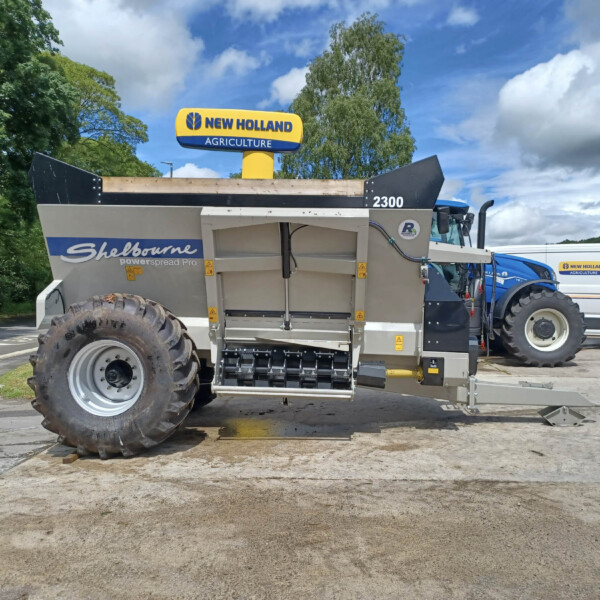 Large agricultural machine with oversized tires in a parking lot; blue sky and trees in the background, a New Holland sign visible on a van in the distance.