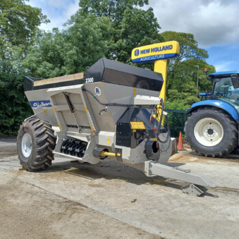 Blue New Holland tractor beside a large gray agricultural machine mounted on a trailer, outdoors with trees in the background.