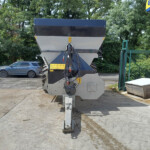 Large hopper-style agricultural spreader mounted on a trailer in an outdoor lot, with a blue SUV nearby and green trees in the background.