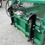 Close-up of a green industrial hydraulic machine with hoses, metal fittings, and a mesh guard behind it.