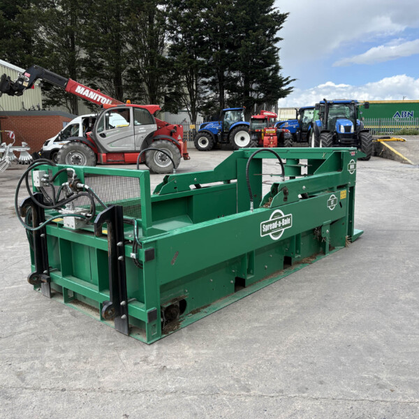 Large green spreader machine labeled 'Spread-a-Bale' in the foreground at an outdoor equipment yard, with blue tractors and a red crane in the background.