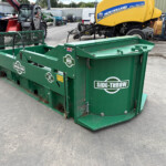 Green Side-Throw waste compactor in an outdoor yard with heavy machinery in background (New Holland yellow machine seen).