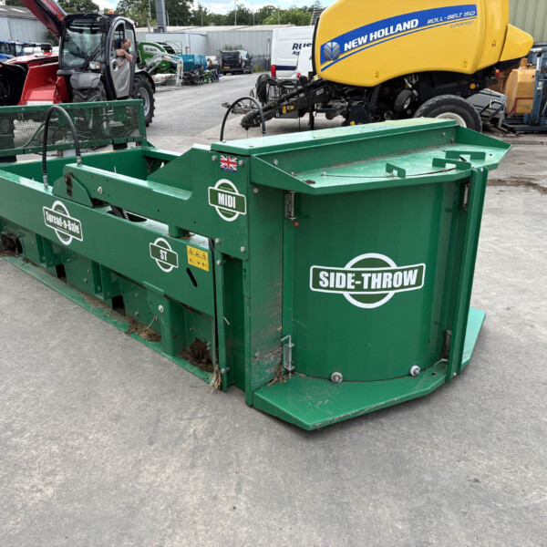 Green Side-Throw waste compactor in an outdoor yard with heavy machinery in background (New Holland yellow machine seen).