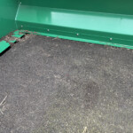 Close-up of the interior corner of a green metal trailer bed with a damaged edge and scattered straw on a dark mat-ish surface