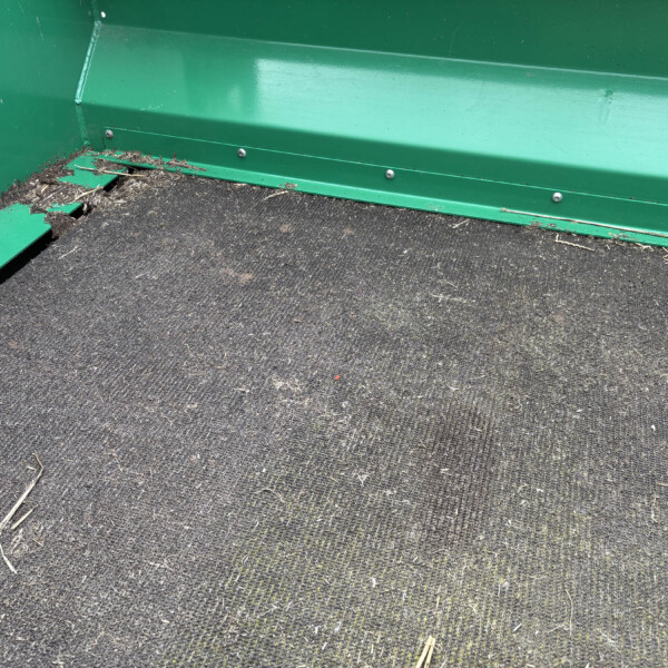 Close-up of the interior corner of a green metal trailer bed with a damaged edge and scattered straw on a dark mat-ish surface