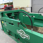 Green bale-wrapping machine in an industrial yard with pallets, stacked tires, and a corrugated metal building in the background.