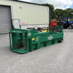 Green agricultural machine in foreground with stakes and a black conveyor, in a yard beside a corrugated metal building.