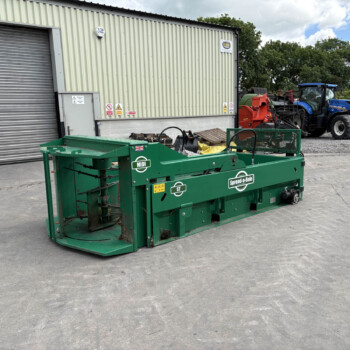 Green agricultural machine in foreground with stakes and a black conveyor, in a yard beside a corrugated metal building.