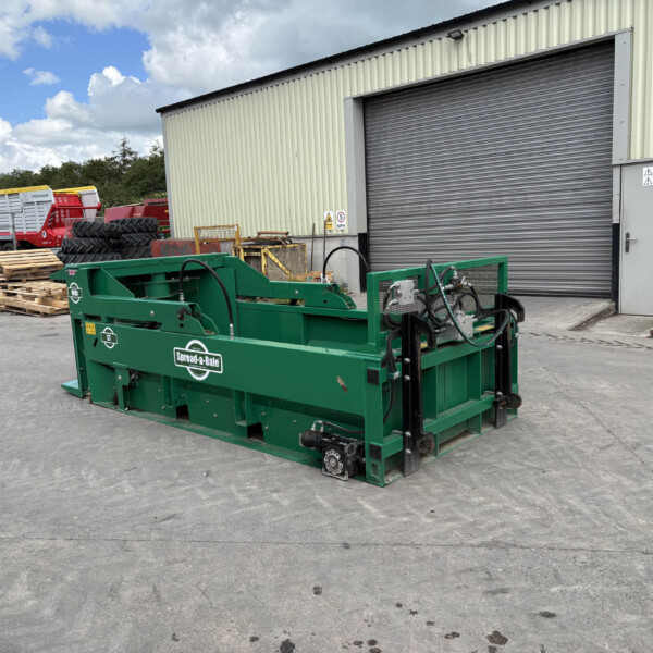 Industrial yard with a large green machinery piece in the foreground and a corrugated metal building with a closed roller shutter in the background.
