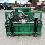 Green industrial machine with hoses and metal frame in a yard, with tractors and tires in the background.