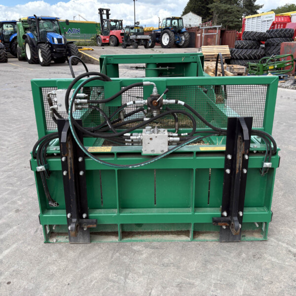Green industrial machine with hoses and metal frame in a yard, with tractors and tires in the background.