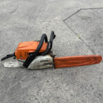 Orange STIHL chainsaw resting on rough concrete pavement with a black handle visible.