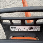 Close-up of a black STIHL equipment housing with an orange bar visible behind it and a manufacturer label nearby.