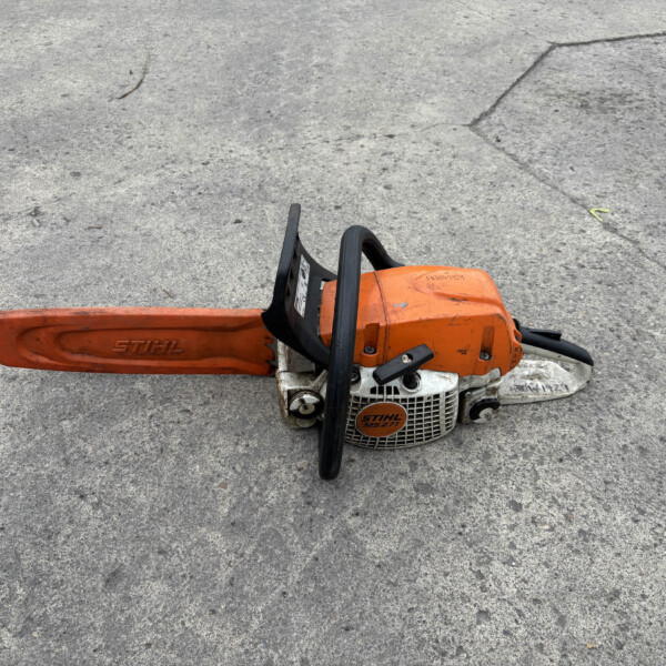 Orange STIHL chainsaw resting on a rough concrete surface, with the long bar extending to the left and black handles visible beneath.