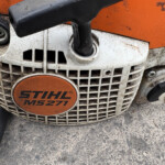 Close-up of an orange and white Stihl MS271 chainsaw, showing the ventilation grille and the round orange MS271 badge with dirt and grime on the housing