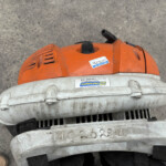 Close-up of an orange handheld leaf blower on a concrete surface, showing a sticker and dusty housing.