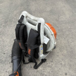 Portable concrete-cutting saw with orange engine housing and black safety straps resting on a gray pavement surface, hose attached to the tool.