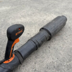 Close-up of a black and orange Stihl garden tool resting on a rough concrete surface, showing the handle and shaft.