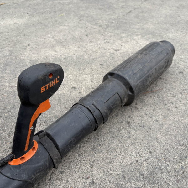 Close-up of a black and orange Stihl garden tool resting on a rough concrete surface, showing the handle and shaft.