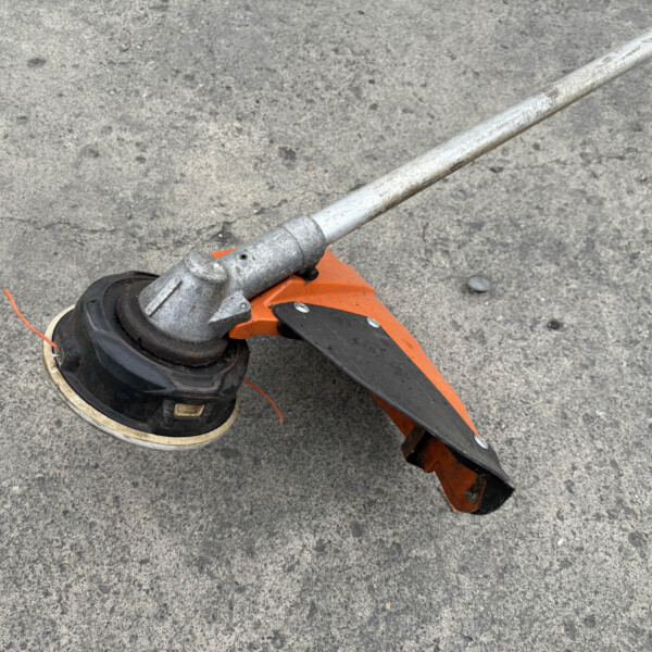 Close-up of an orange and black string trimmer on rough asphalt, showing the metal shaft and cutting guard from above.