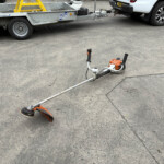 Trailer hitched to a white pickup at a worksite; an orange STIHL power tool lies on the asphalt in the foreground.