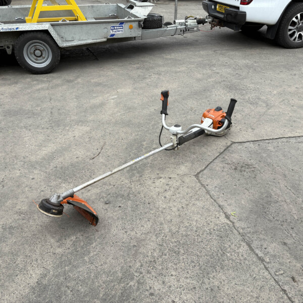 Trailer hitched to a white pickup at a worksite; an orange STIHL power tool lies on the asphalt in the foreground.