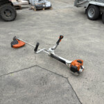Orange and white string trimmer lying on its side on a concrete lot, with trucks and pallets in an industrial yard behind it.