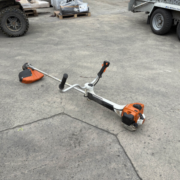 Orange and white string trimmer lying on its side on a concrete lot, with trucks and pallets in an industrial yard behind it.