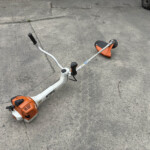Orange STIHL gas-powered string trimmer lying on a cracked concrete driveway, with handlebars and cutting head visible.