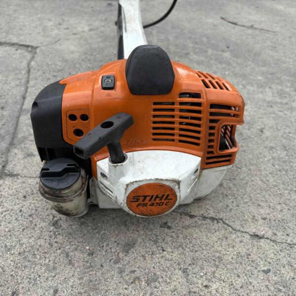 Close-up of an orange STIHL FS 410 C gas trimmer sitting on a rough concrete surface, with black handle grip visible on the left.
