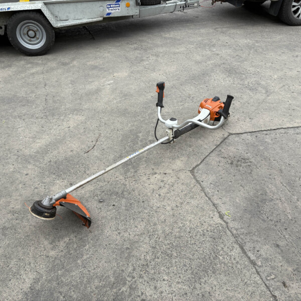 Close-up of a long orange power tool lying on gray asphalt, with a circular blade guard at the end and a trailer visible in the background.