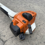 Close-up of an orange gas-powered chainsaw resting on a rough concrete surface, showing the housing, vents, and black handle grip.