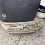 Close-up of a dirty lawn tool's rear housing with a worn STIHL label and a foot pedal, showing orange latch on the side.