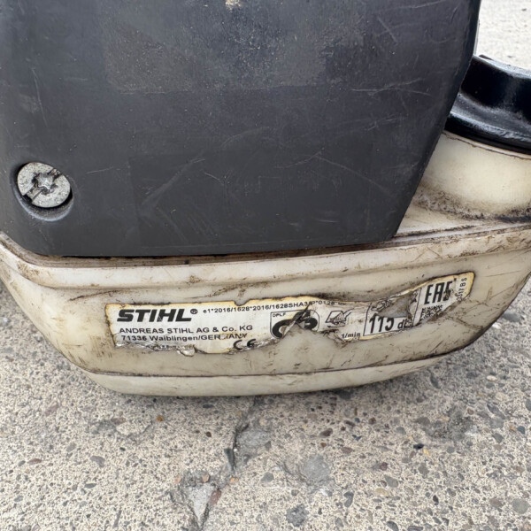 Close-up of a dirty lawn tool's rear housing with a worn STIHL label and a foot pedal, showing orange latch on the side.