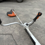 Lawn trimmer with an orange guard lying on a concrete driveway, handle in the foreground.